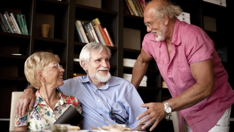 Senior caucasian couple sitting at a table. The man has his arm around his wife. There is another senior aged man standing next to them smiling and talking to them.